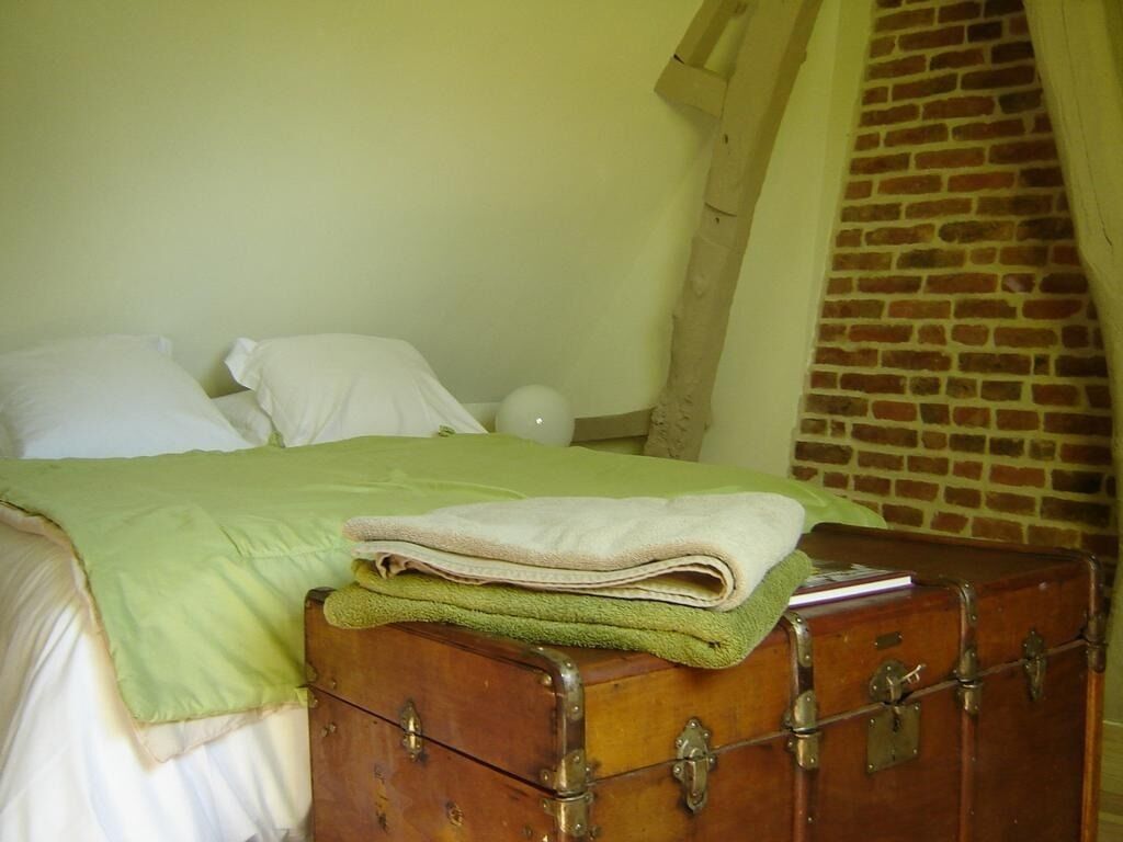 Una cama con una manta y almohadas verdes, un baúl de madera con una toalla enrollada, y una pared de ladrillo al fondo.