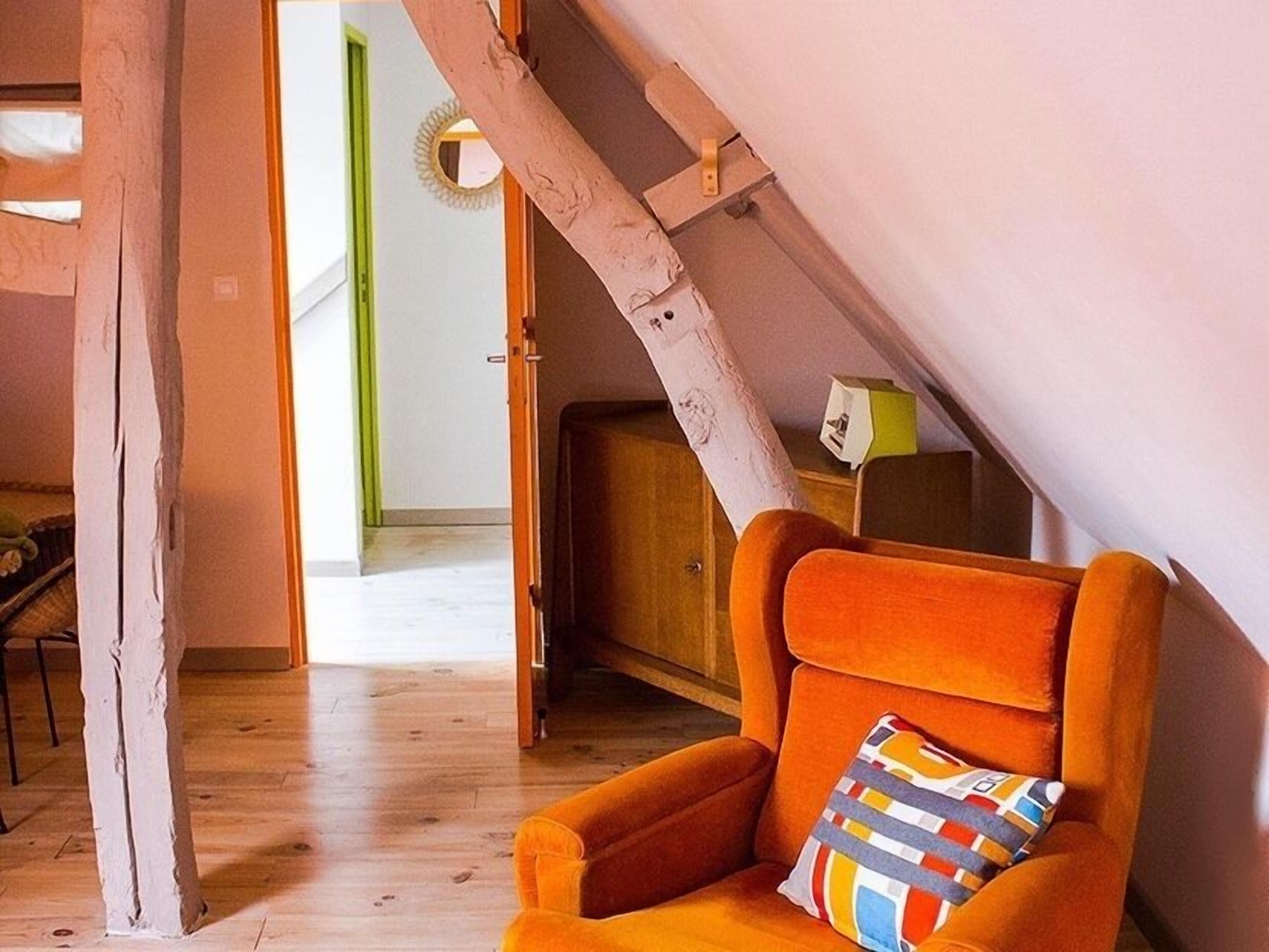 Een gezellige kamer met een houten vloer, een levendige oranje fauteuil en een houten kast.