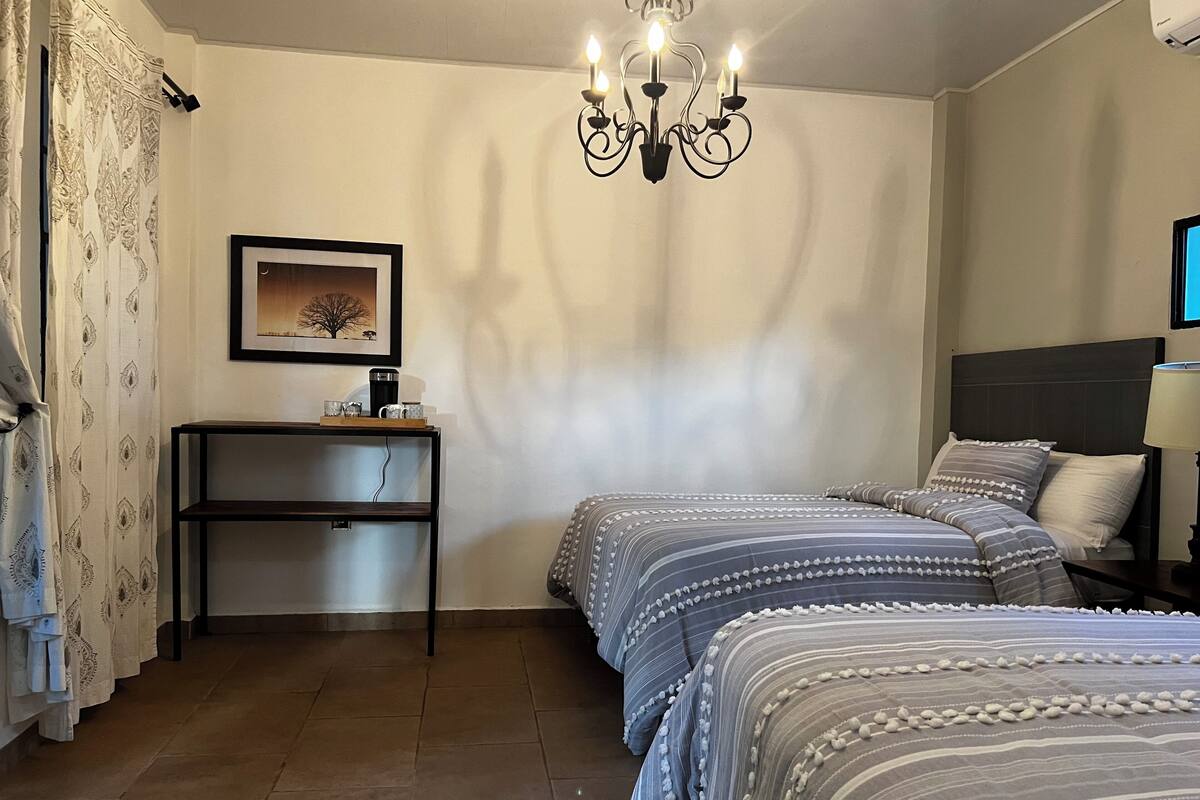 Un dormitorio con dos camas, una mesita de noche, un escritorio, una silla, una araña y un cuadro enmarcado en la pared.