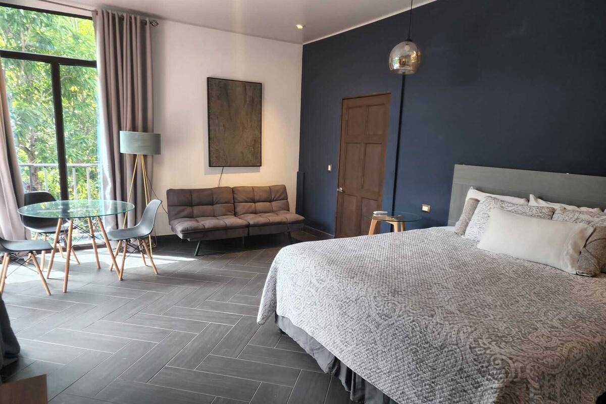 Un dormitorio moderno con una cama grande, un sofá, una mesa de vidrio y vistas al verde exterior.