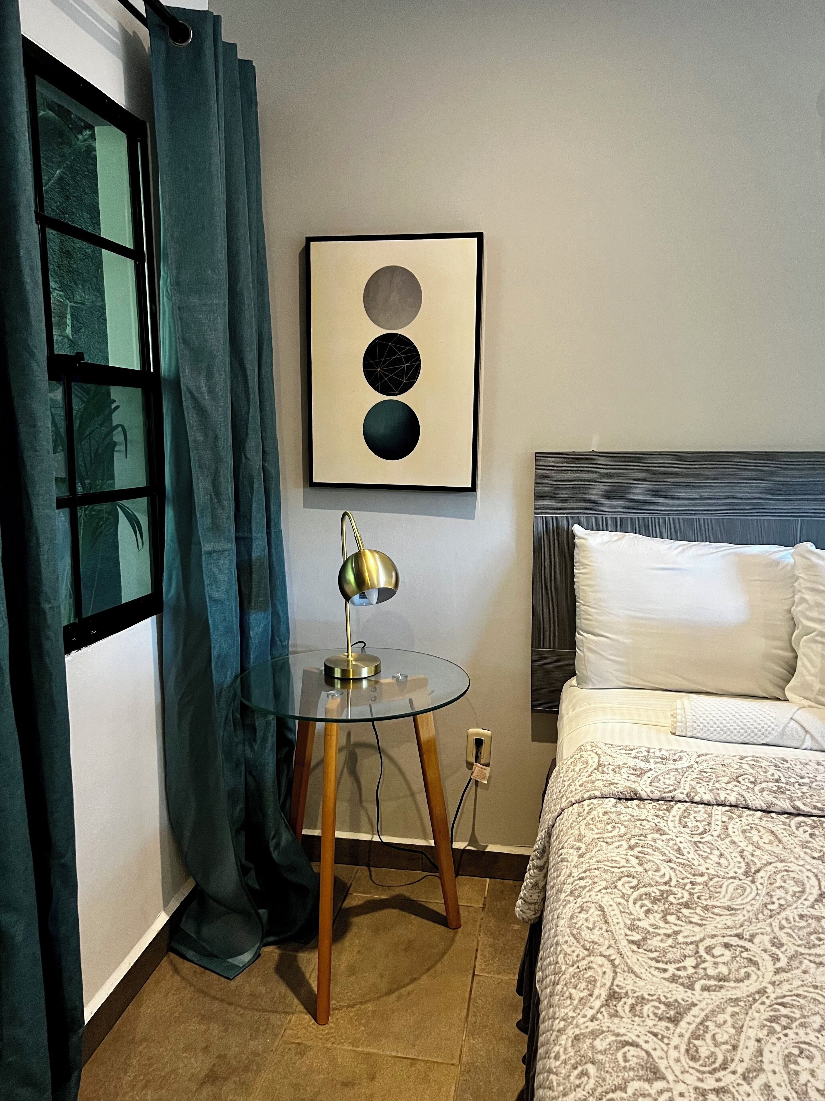 Habitación de hotel con cama, una mesita de vidrio con lámpara, una obra de arte enmarcada y un ventanal con cortinas.