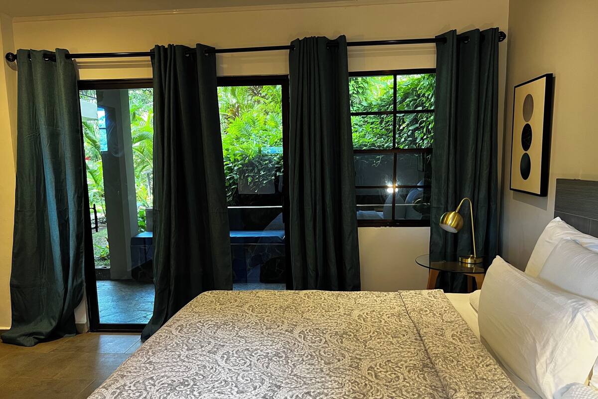 Habitación de hotel con cama, mesita de noche con lámpara, ventana con cortinas y vista a zonas verdes.