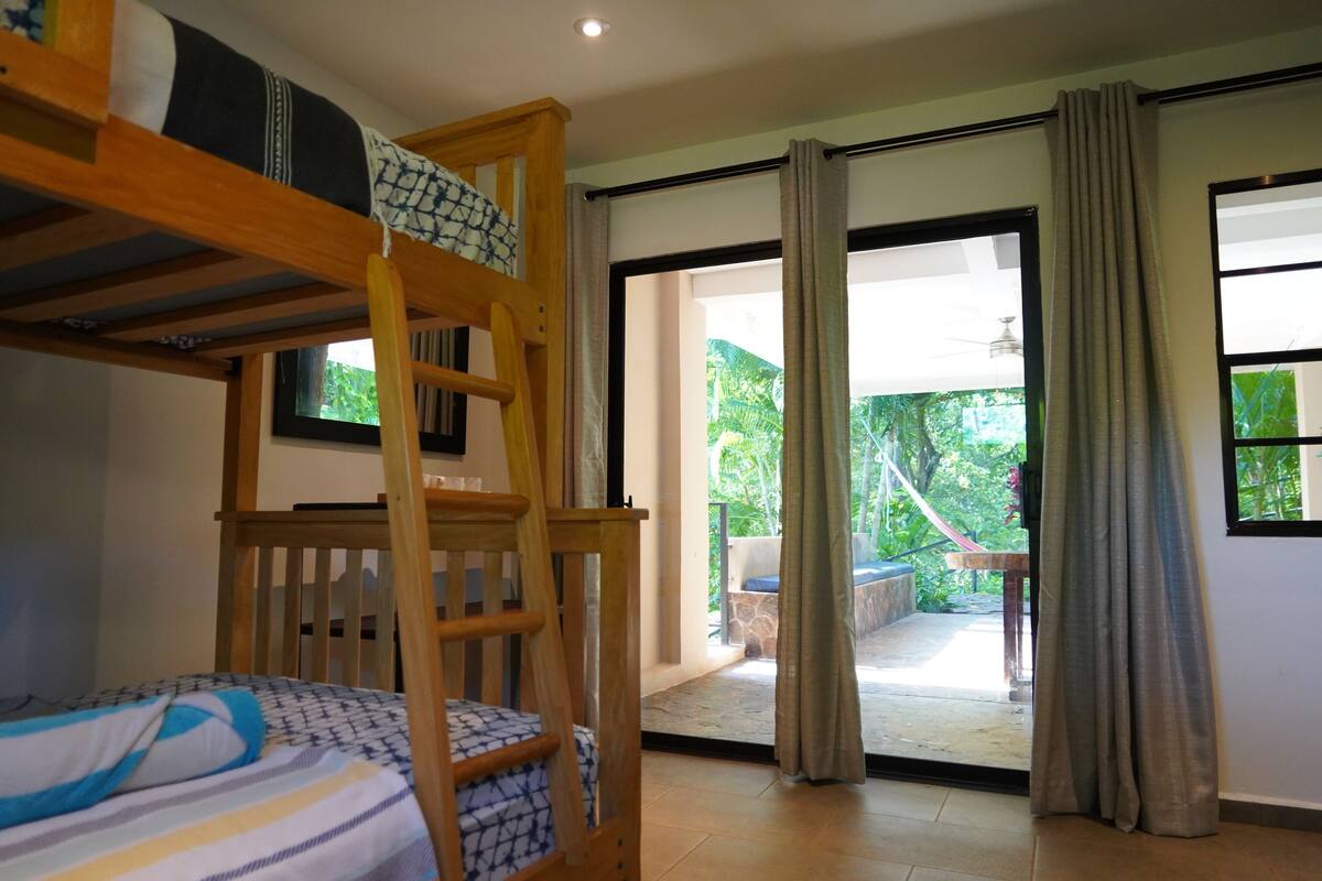 Una habitación con literas, una escalera de madera, una colcha estampada y vista a un espacio exterior a través de un ventanal.