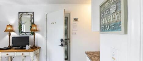 A room with a white door, a mirror, a desk with a computer, two lamps, and a sign that says "The Beach is my Happy Place."
