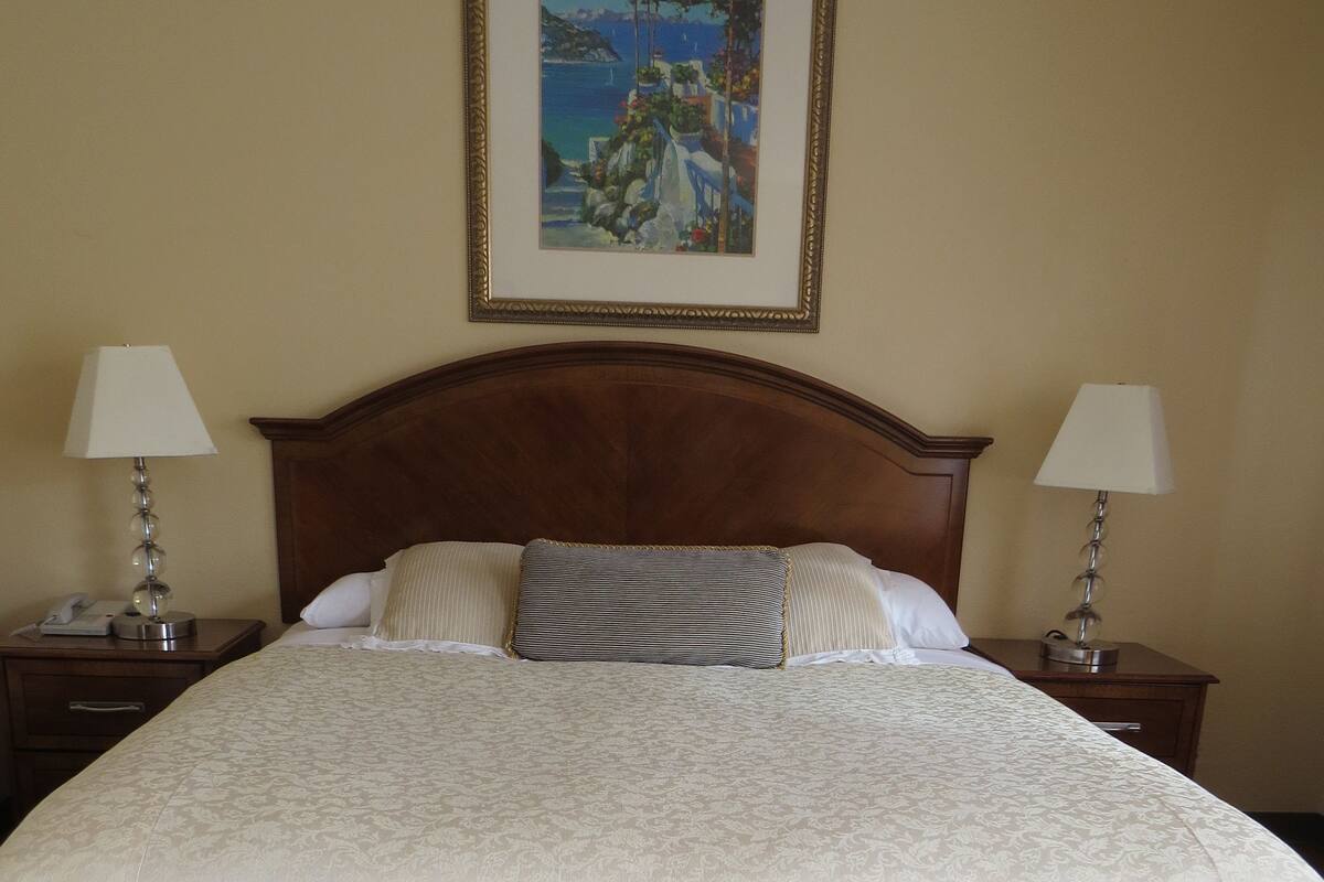A neatly made bed with a white bedspread and striped pillows, a wooden headboard, two bedside lamps, and a framed painting above the bed.