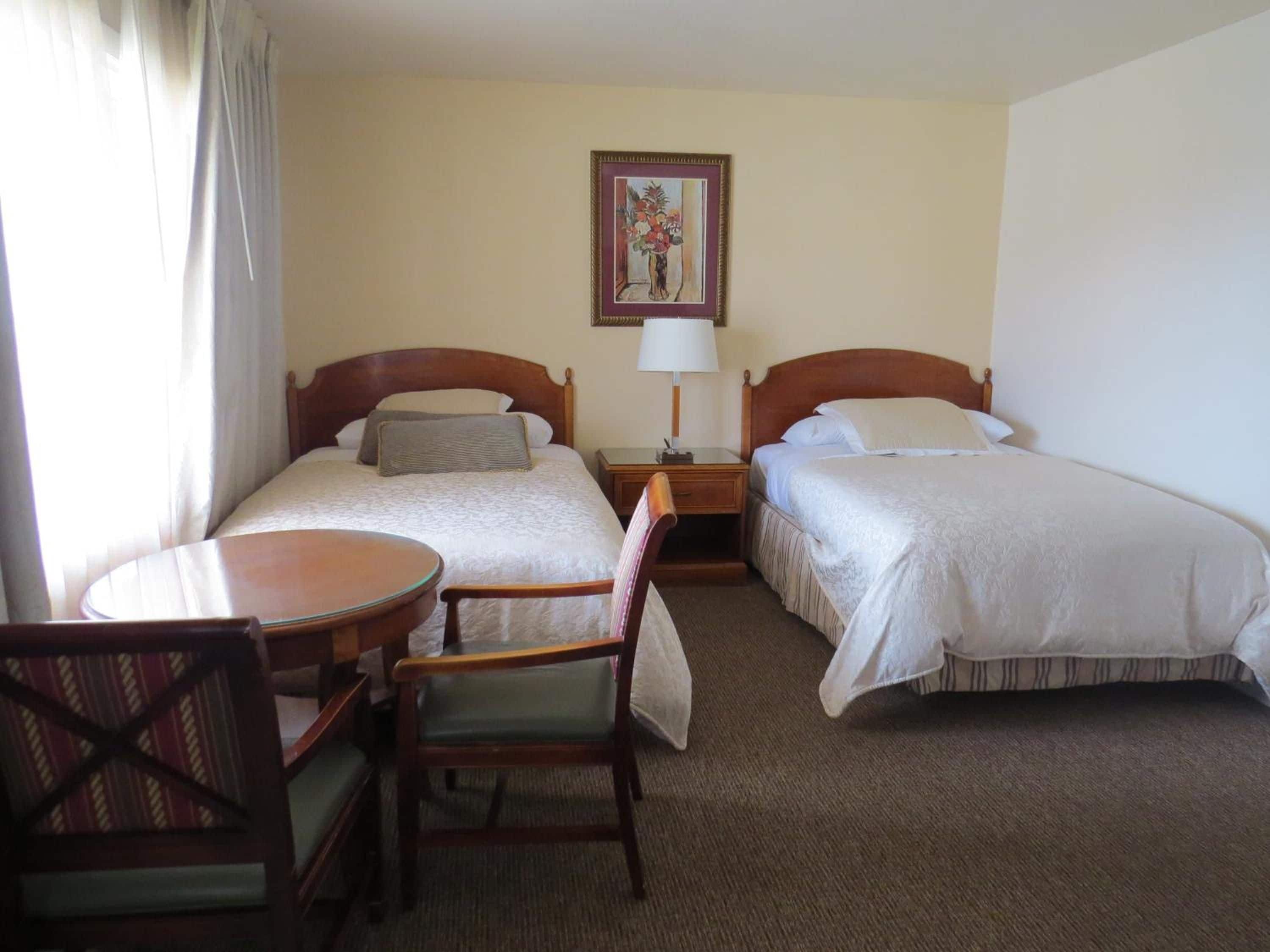 A hotel room with two beds, a small table, a chair, and a painting on the wall.