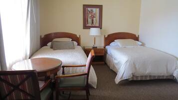 A hotel room with two beds, a small table, a chair, and a painting on the wall.