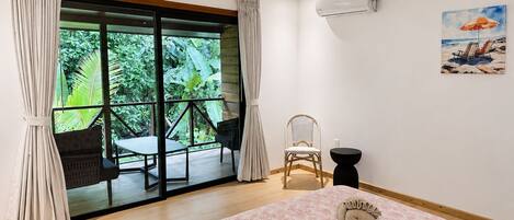 A bedroom with a large bed, a chair, a small table, and a view of greenery through a sliding door.