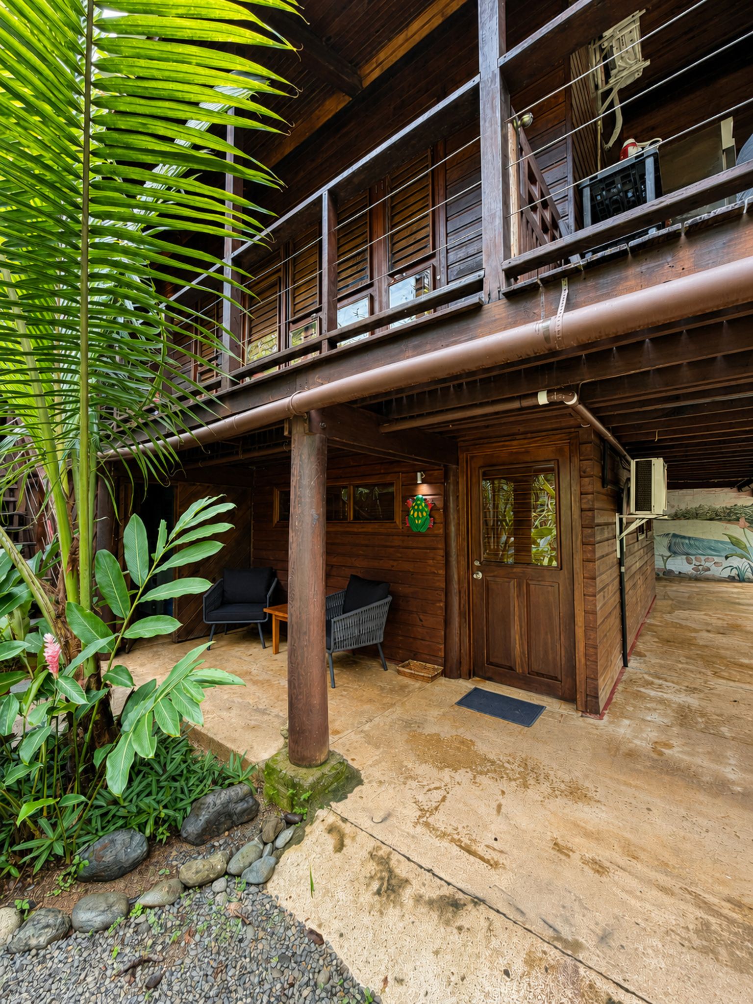 Una cabaña de madera con balcón, un porche con asientos y un jardín con plantas tropicales.