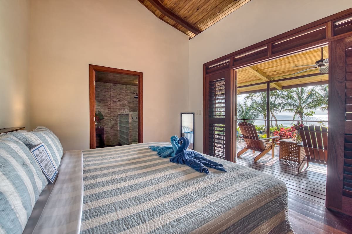 A hotel room with a bed, a wooden door leading to a balcony, and a view of the ocean.