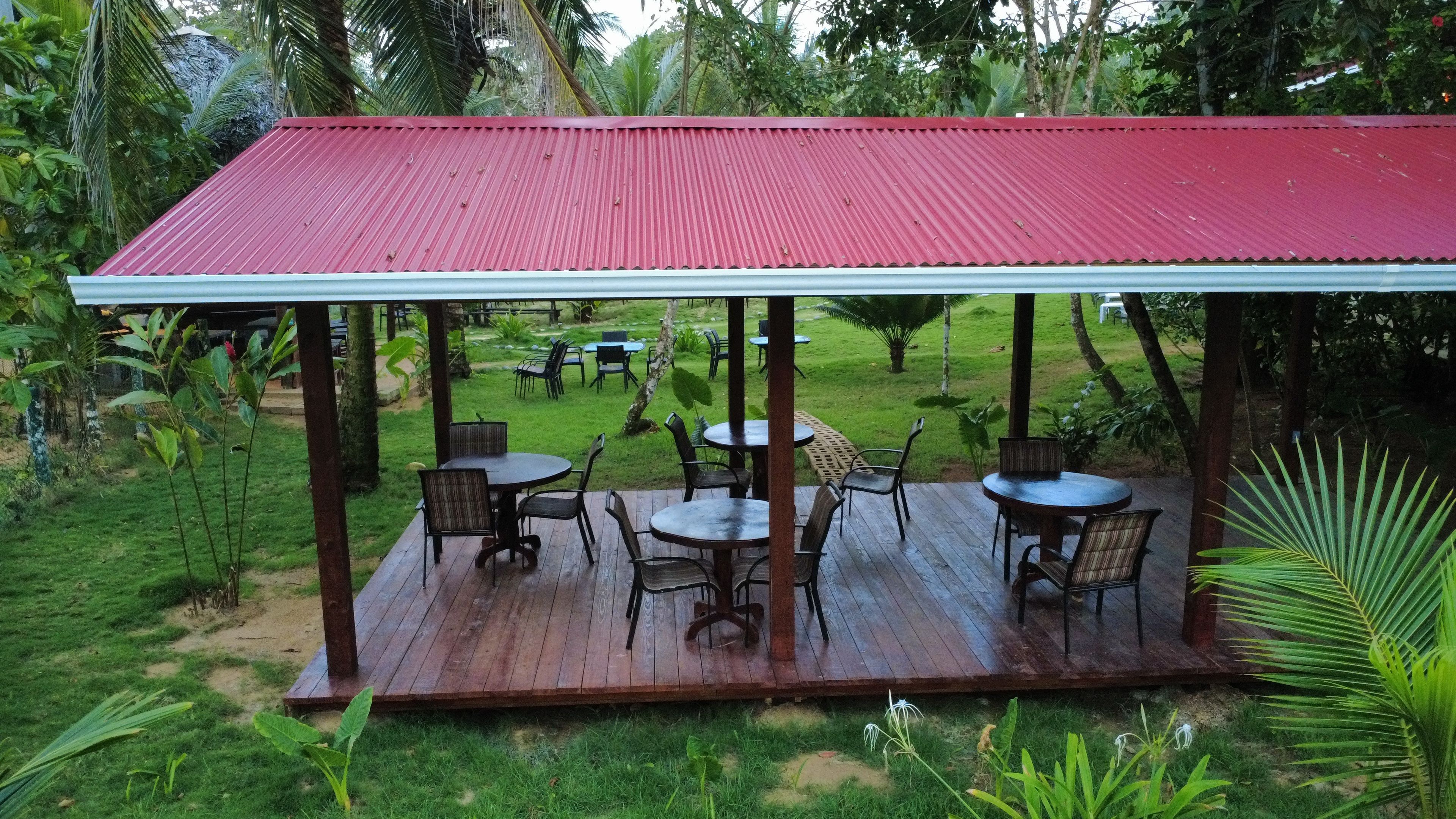 Un patio exterior con techo rojo, rodeado de follaje y amueblado con mesas y sillas.