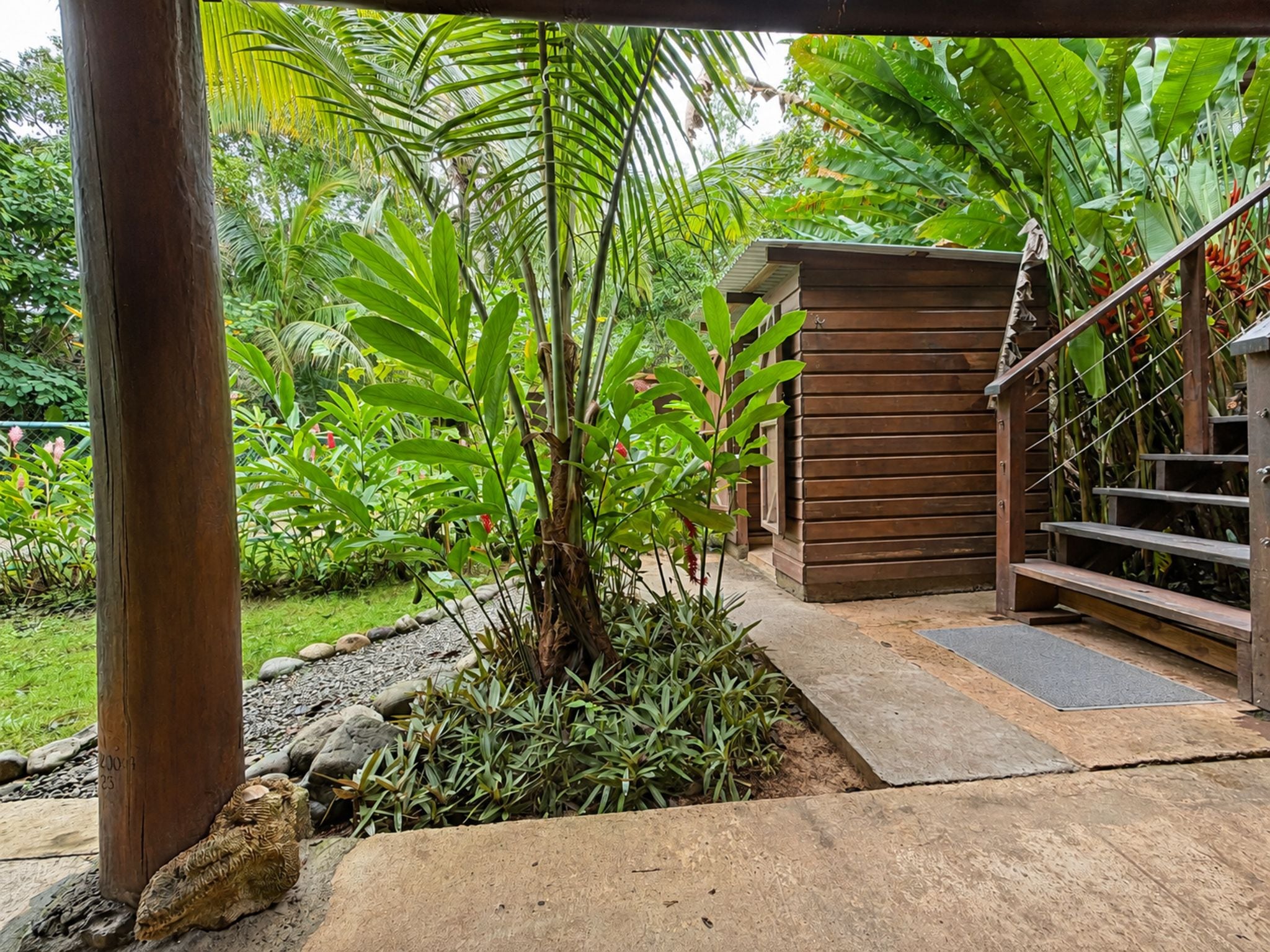 A wooden staircase leading to a small wooden structure, surrounded by lush greenery and a garden area with various plants and a stone pathway.