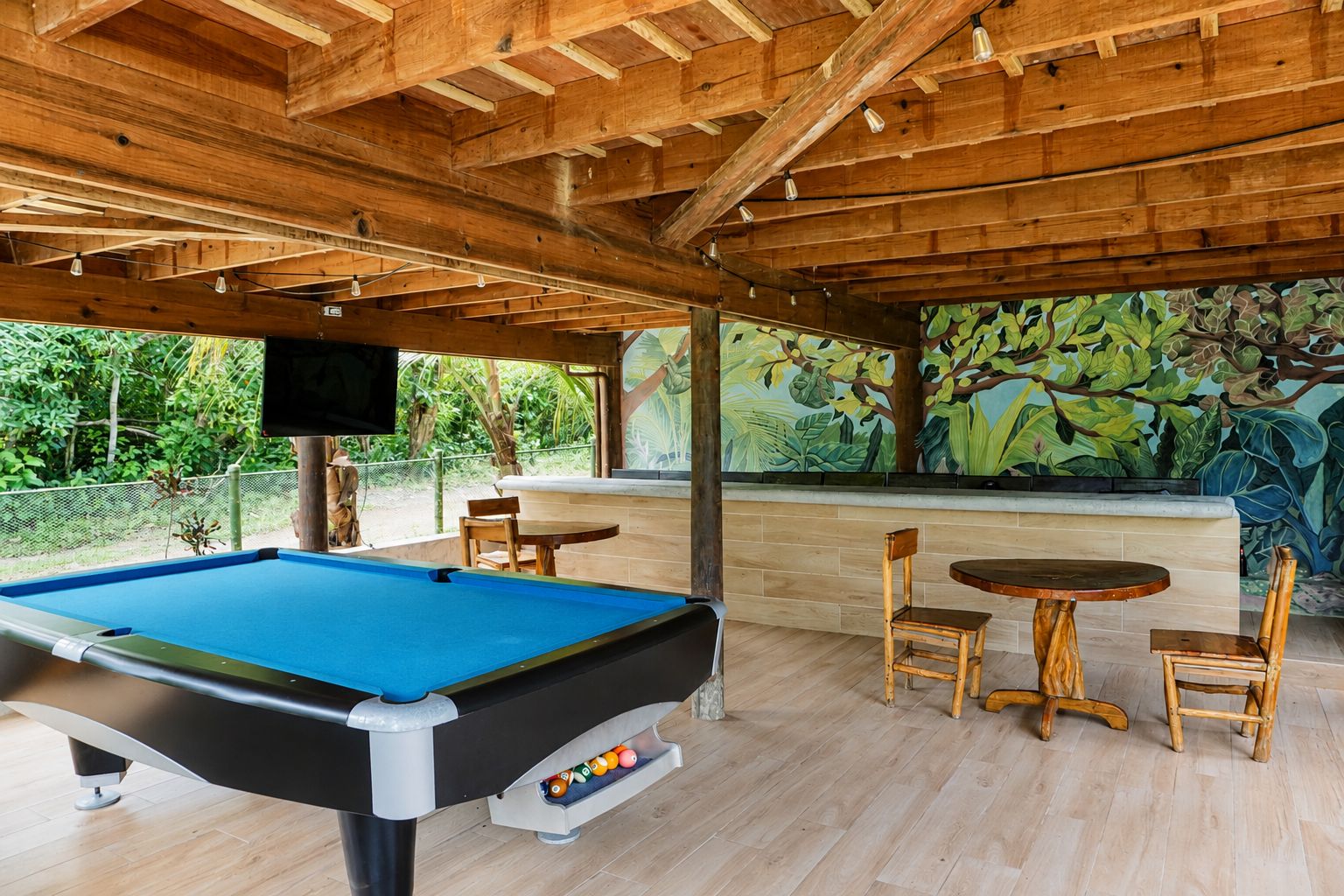 Una mesa de billar con sillas, un televisor y un mural de follaje tropical.