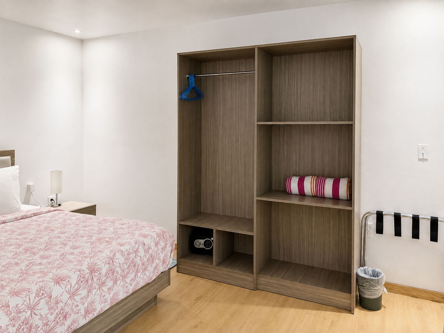 A bedroom with a bed, a wardrobe with multiple shelves, a hanging rack, and a small trash bin.