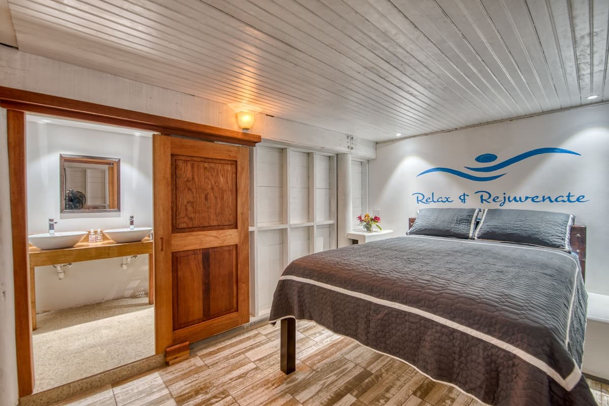 A bedroom with a bed, a wooden wardrobe, a bathroom with a sink and mirror, and a wall with a logo and text.