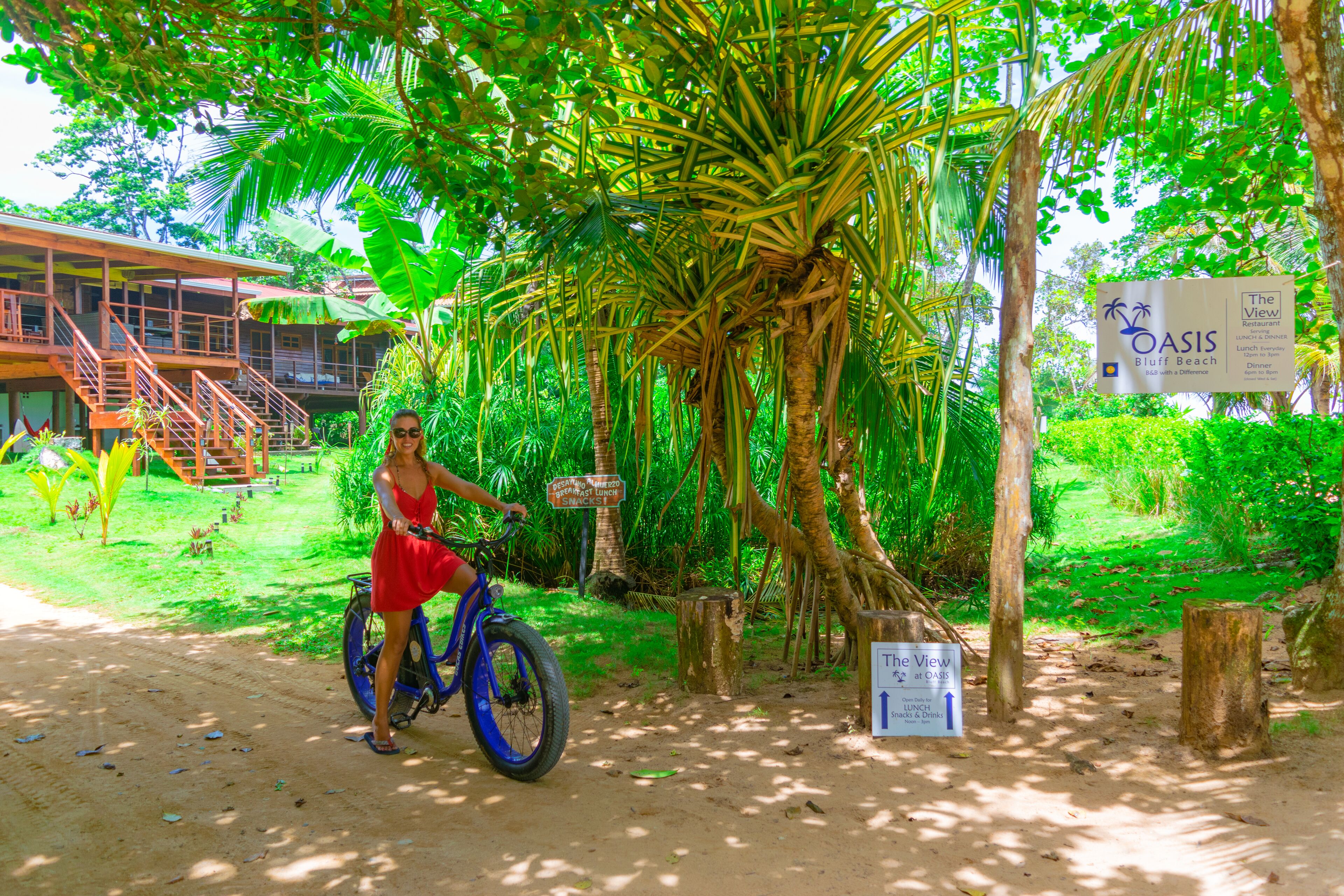 Una mujer con un vestido rojo montando una bicicleta azul sobre un sendero de tierra, rodeada de exuberante vegetación y palmeras.