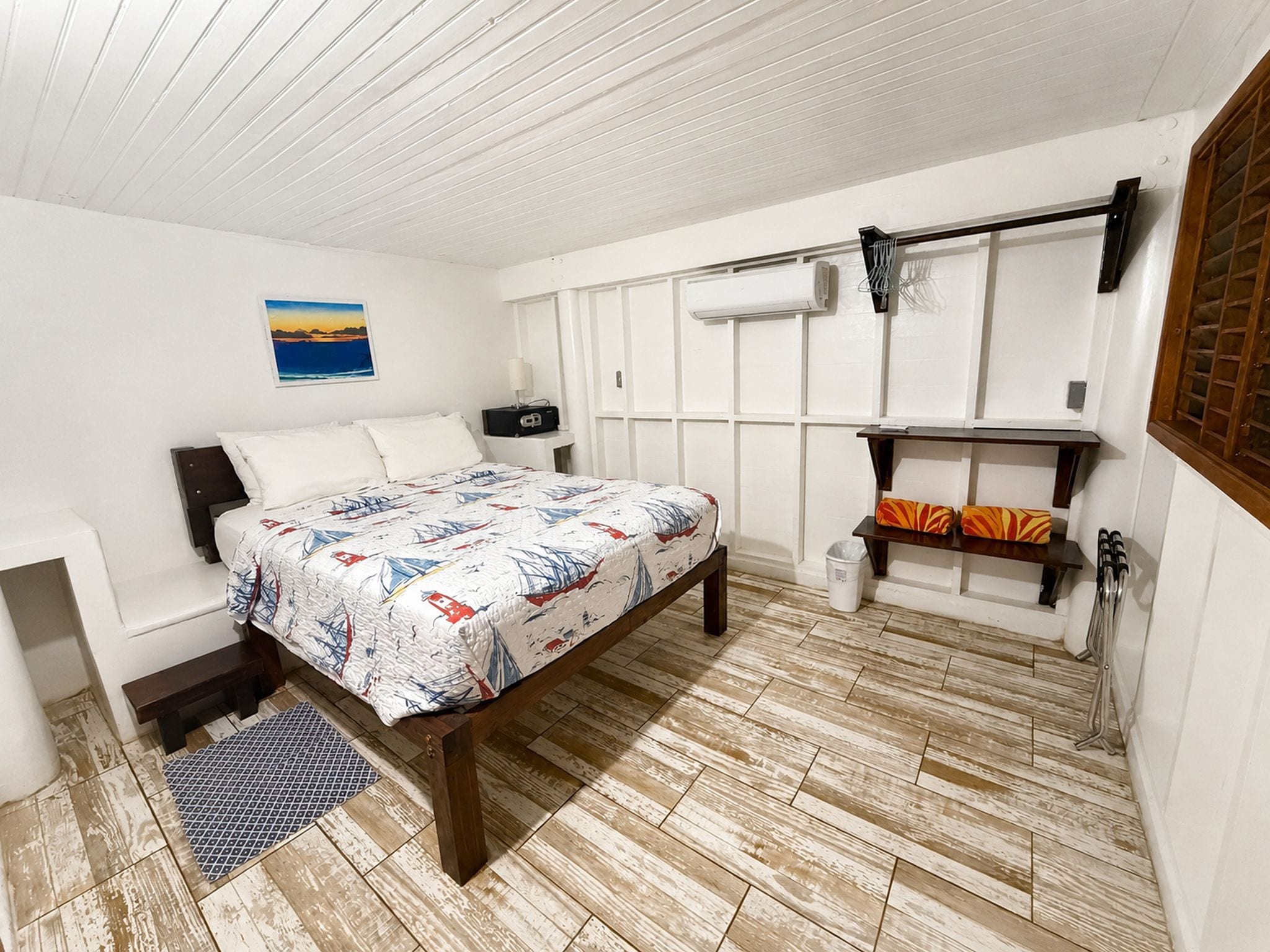 A bedroom with a bed, a desk, a chair, a radiator, a shelf with towels, and a picture on the wall.