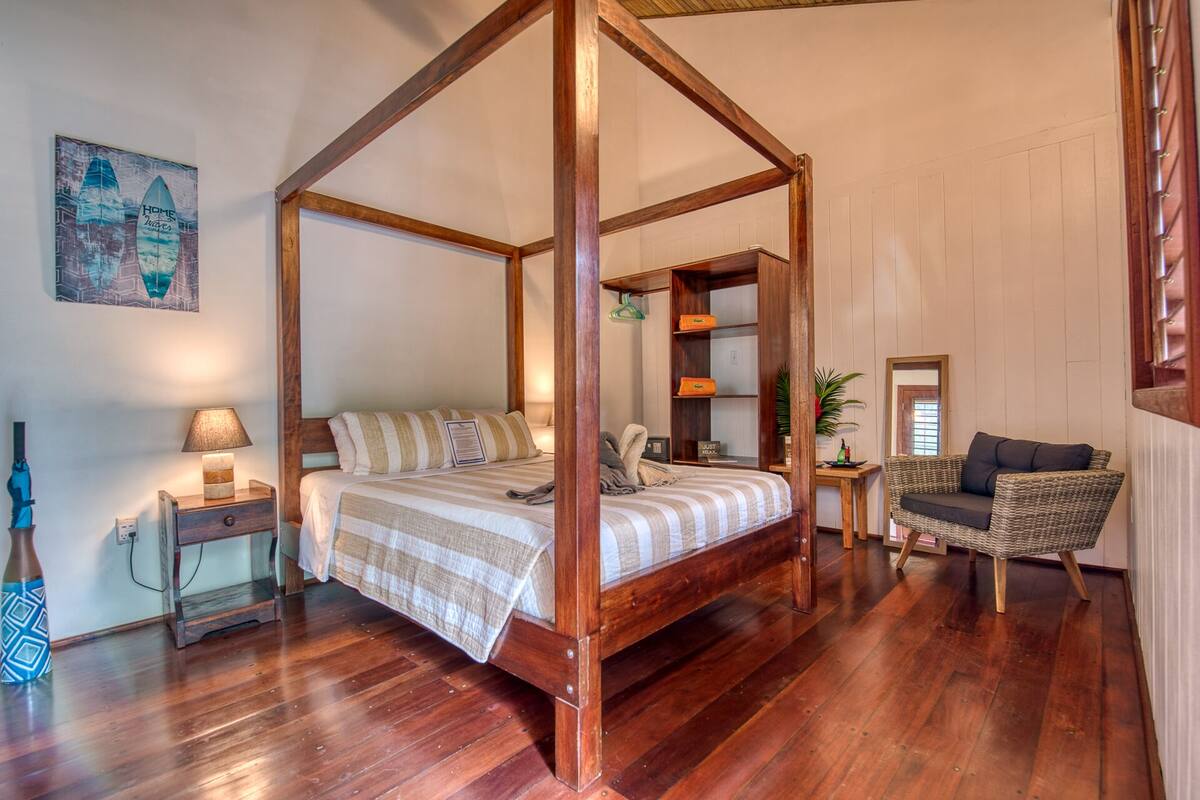 A four-poster bed with a striped bedspread, a bedside table with a lamp, a wicker armchair, and a wooden shelf unit with books and decorative items.