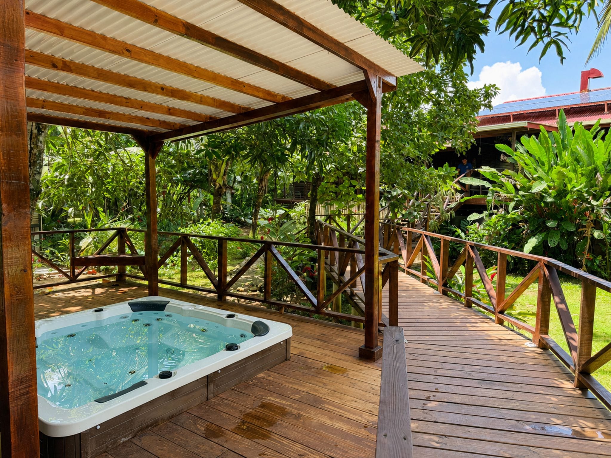 A wooden deck with a hot tub, surrounded by lush greenery and a covered area with a metal roof.