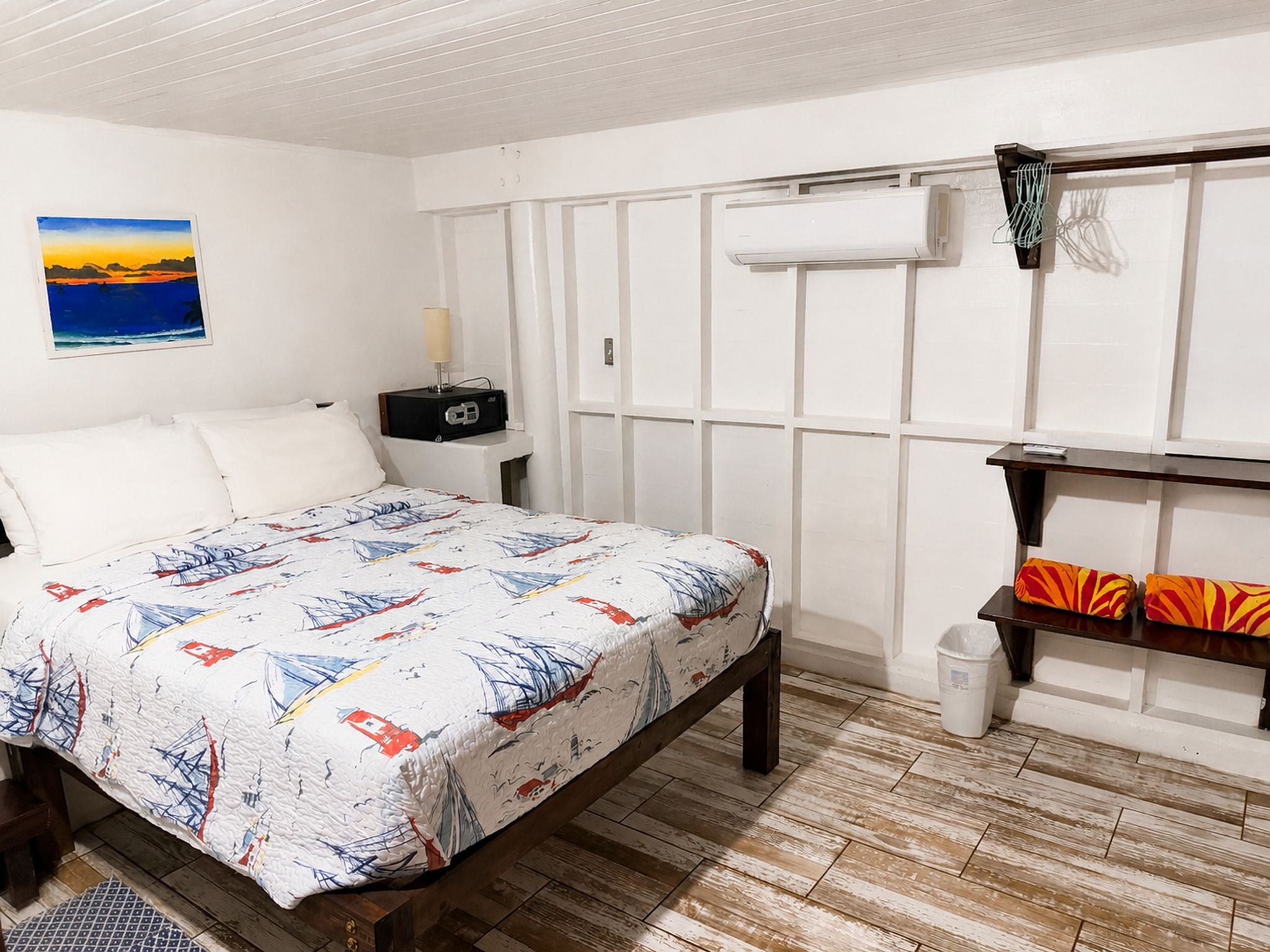 Un dormitorio con cama, mesita de noche, lámpara, aire acondicionado instalado en la pared y estante con cojines.