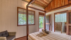 A bedroom with a bed, a wicker sofa, wooden beams, and a view of greenery through the windows. - Oasis Bluff Beach (Bocas del Toro)