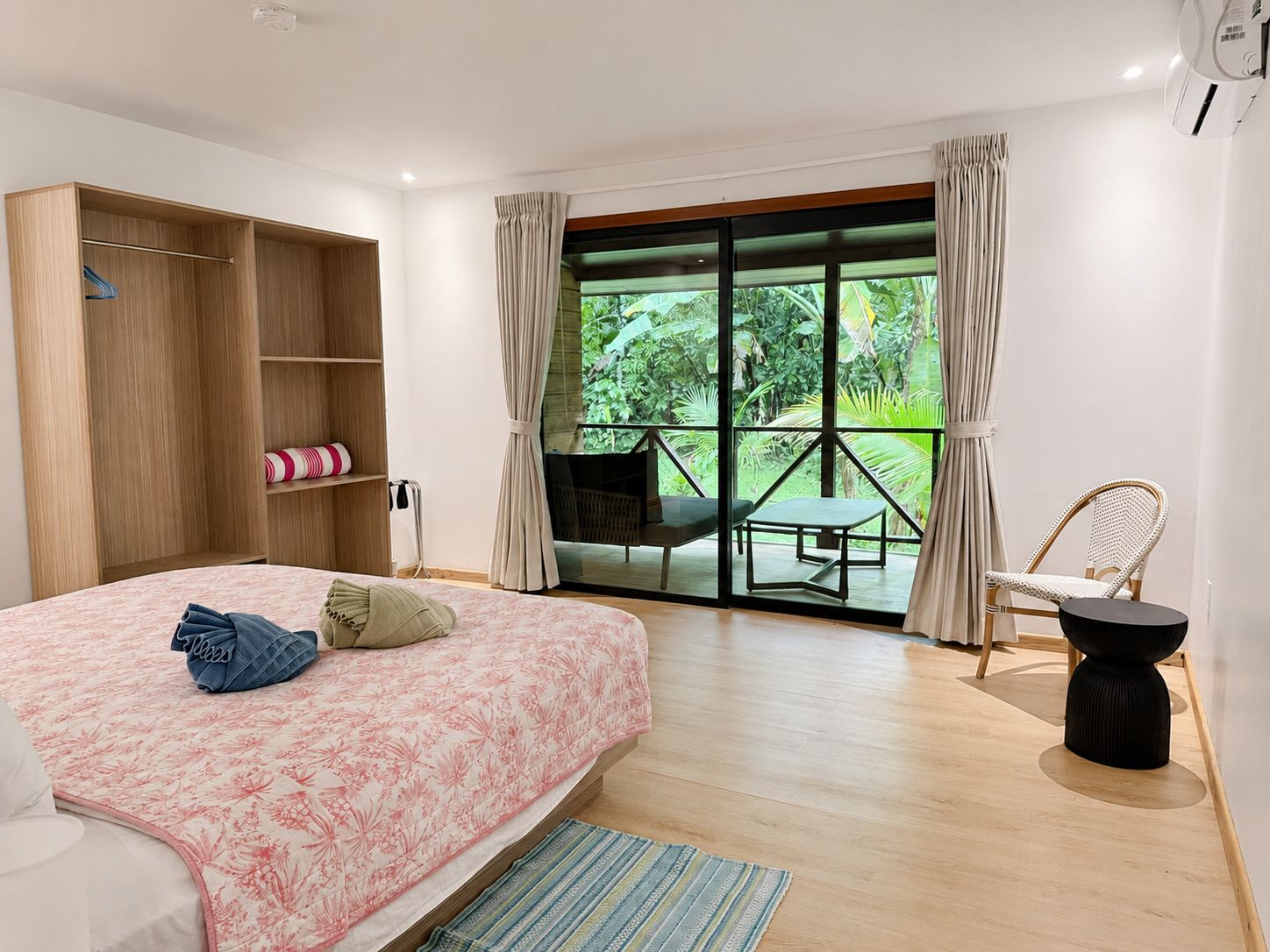 A bedroom with a bed, a wardrobe, a small table, a chair, and a view of greenery through a sliding glass door.