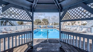 A wooden deck leading to a pool with a view of buildings and palm trees. - The Cove 217a 1 Bedroom Condo by RedAwning (Gulf Shores)
