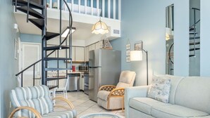 A modern living room with a sofa, two armchairs, a glass coffee table, a kitchen area with a refrigerator, and a staircase leading to an upper level. - The Cove 217a 1 Bedroom Condo by RedAwning (Gulf Shores)