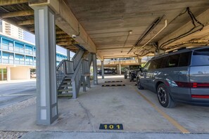 A covered parking area with a car parked in the foreground, a staircase to an upper level, and a building with blue railings in the background. - Sandy Shores West 101 2 Bedroom Condo by RedAwning (Gulf Shores)