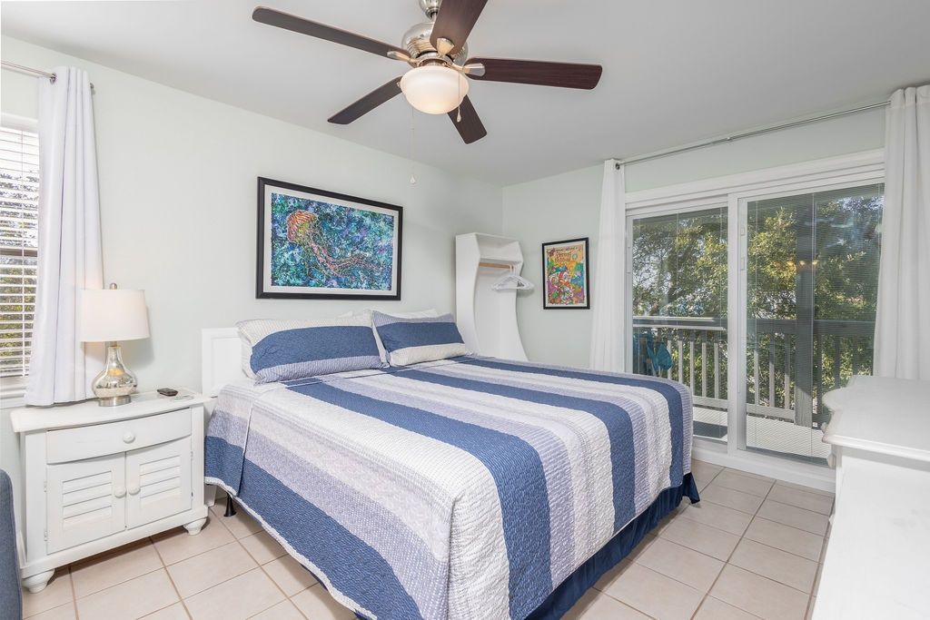 Quarto com cama, mesa de cabeceira, ventoinha no teto e janela com vista para o exterior.