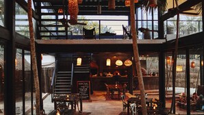 A restaurant with wooden tables and chairs, hanging wicker lamps, and large windows.