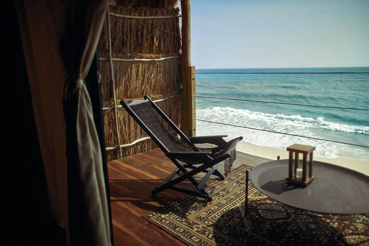 Habitación frente a la playa con vista al océano, pared de paja, mesa de madera y una silla de lounge.