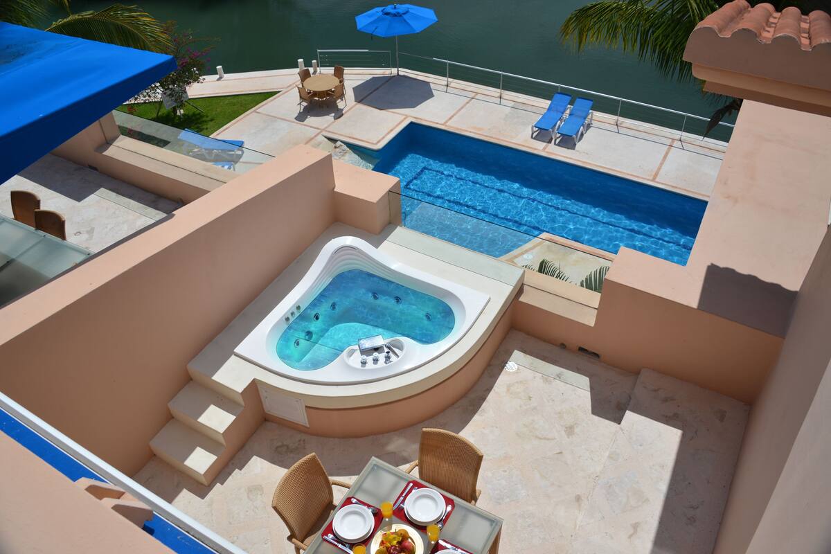 A rooftop pool area with a hot tub, lounge chairs, and a dining table with breakfast items.