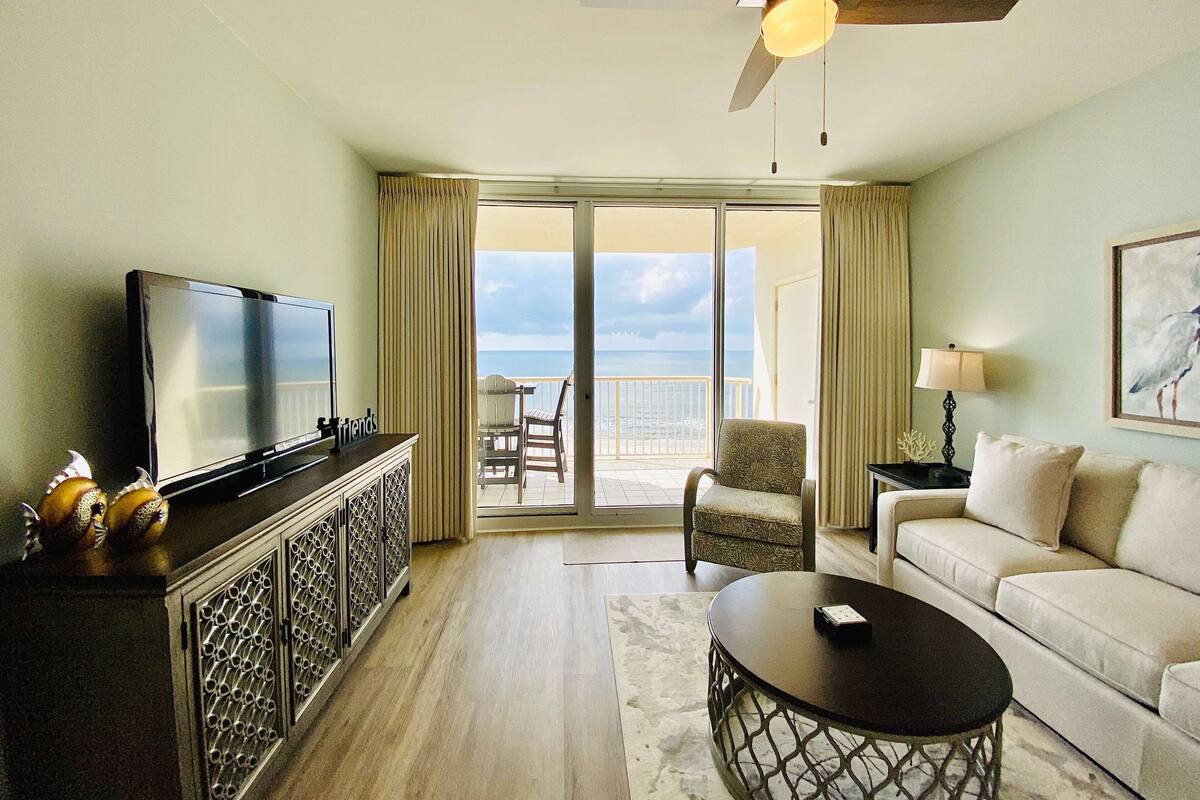 A hotel room with a television, a ceiling fan, a balcony with a view of the sea, a sofa, and a coffee table.