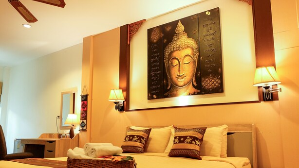 A hotel room with a large Buddha painting on the wall, a bed with patterned pillows, a bedside table with a lamp, and a wicker basket with towels. - QG Resort (Bangkok)
