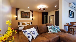 A well-lit room with a brown sofa, a bed with white and brown bedding, a painting on the wall, and a dining area with a table and chairs. - QG Resort (Bangkok)
