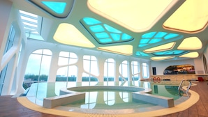 Et rummeligt poolområde indendørs med et centralt boblebad, store vinduer og et moderne loftdesign.