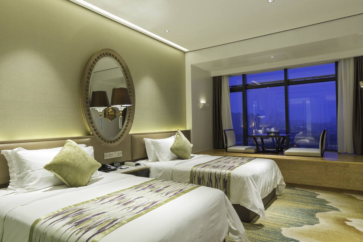 A hotel room with two beds, a large mirror, a view of the city at night, and a seating area with a table and chairs.
