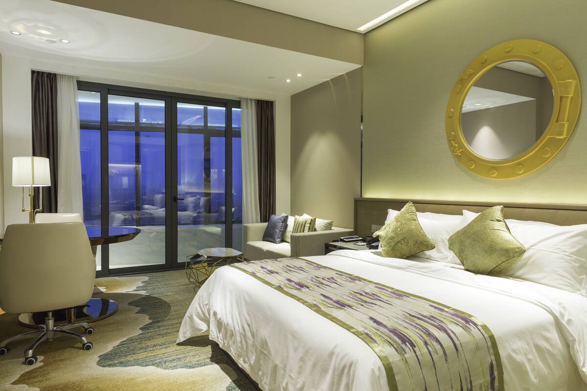 A hotel room with a large bed, a desk, a chair, and a view of the city through large windows.