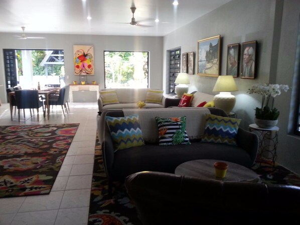 A living room with a sofa, armchairs, a coffee table, and a dining area with a table and chairs. There are paintings on the walls and a plant on a side table.
