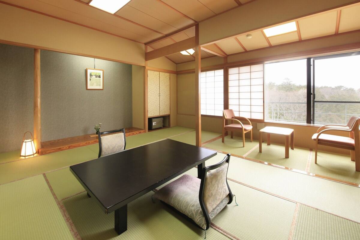 Une pièce avec un sol recouvert de tatamis, une petite table, des chaises, un meuble et une fenêtre.