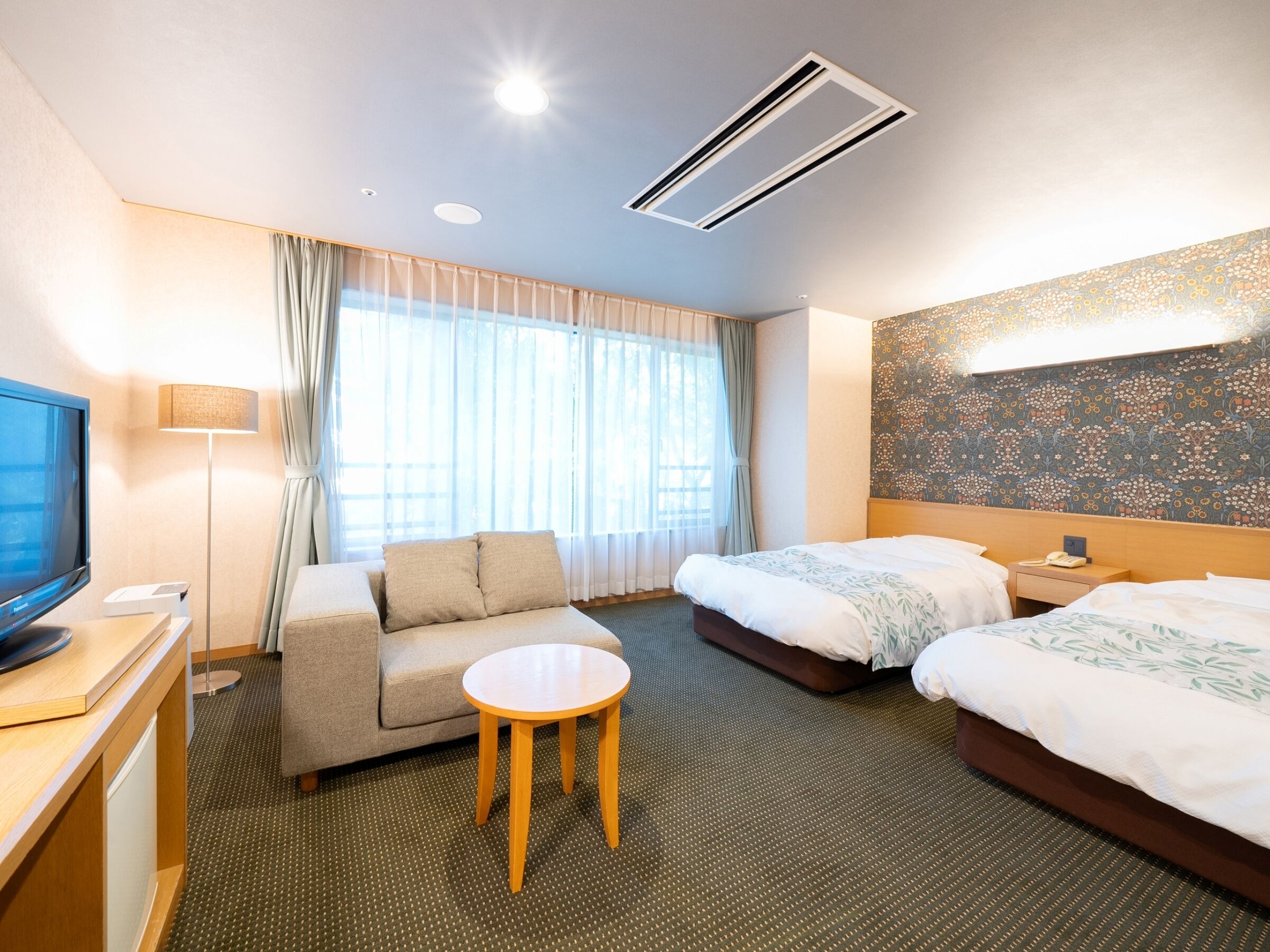 Une chambre d’hôtel avec un canapé, une petite table ronde, deux lits, une télévision et un mur décoré d’un motif.