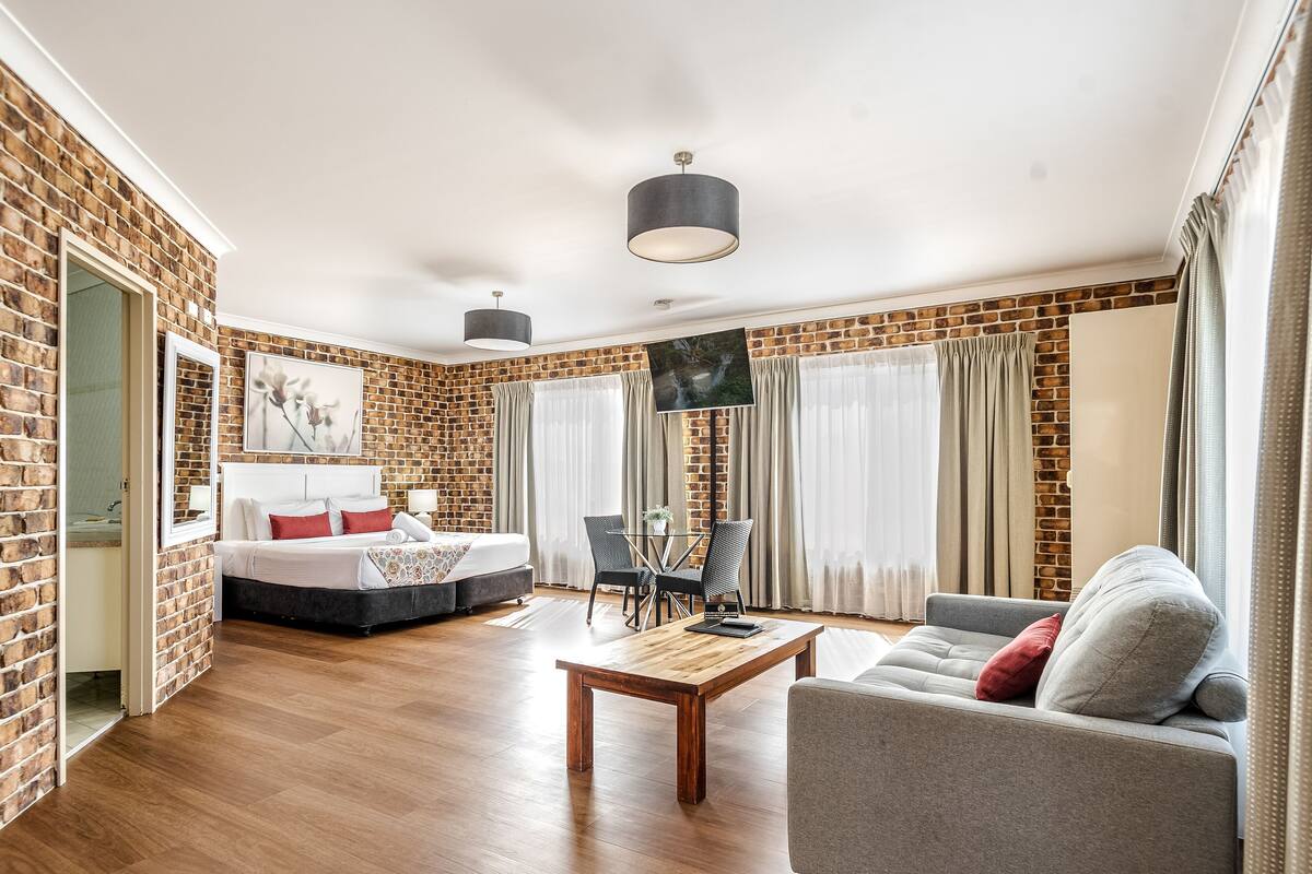 A modern hotel room with a brick wall, a bed, a sofa, a coffee table, and a TV.