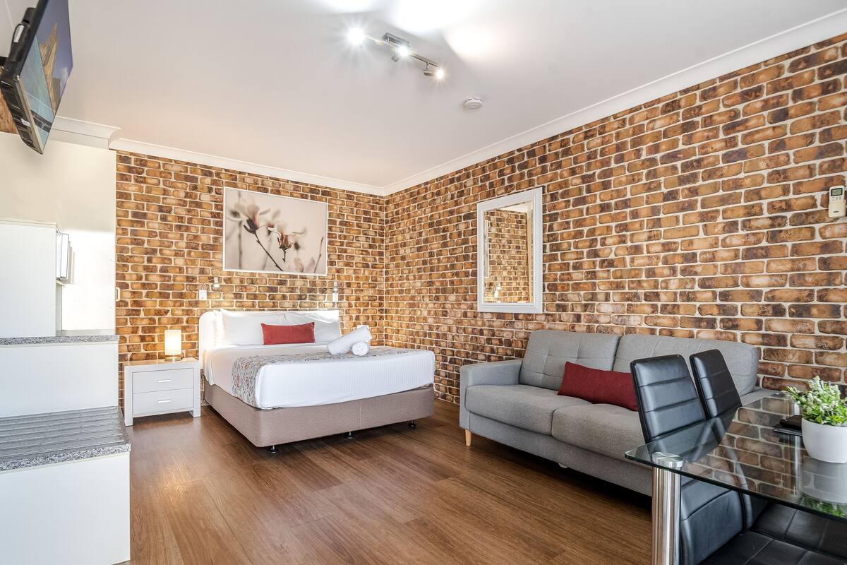 A modern bedroom with a brick-patterned wall, a bed, a grey sofa, and a glass table.