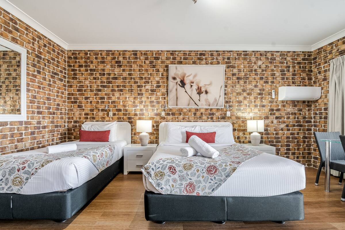 A hotel room with two beds, a brick-patterned wall, a framed floral artwork, bedside lamps, and a chair.