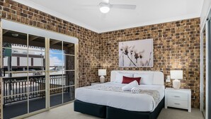 Een slaapkamer met een bakstenen muur, een bed met wit beddengoed, een nachtkastje en een raam met uitzicht op een gebouw.