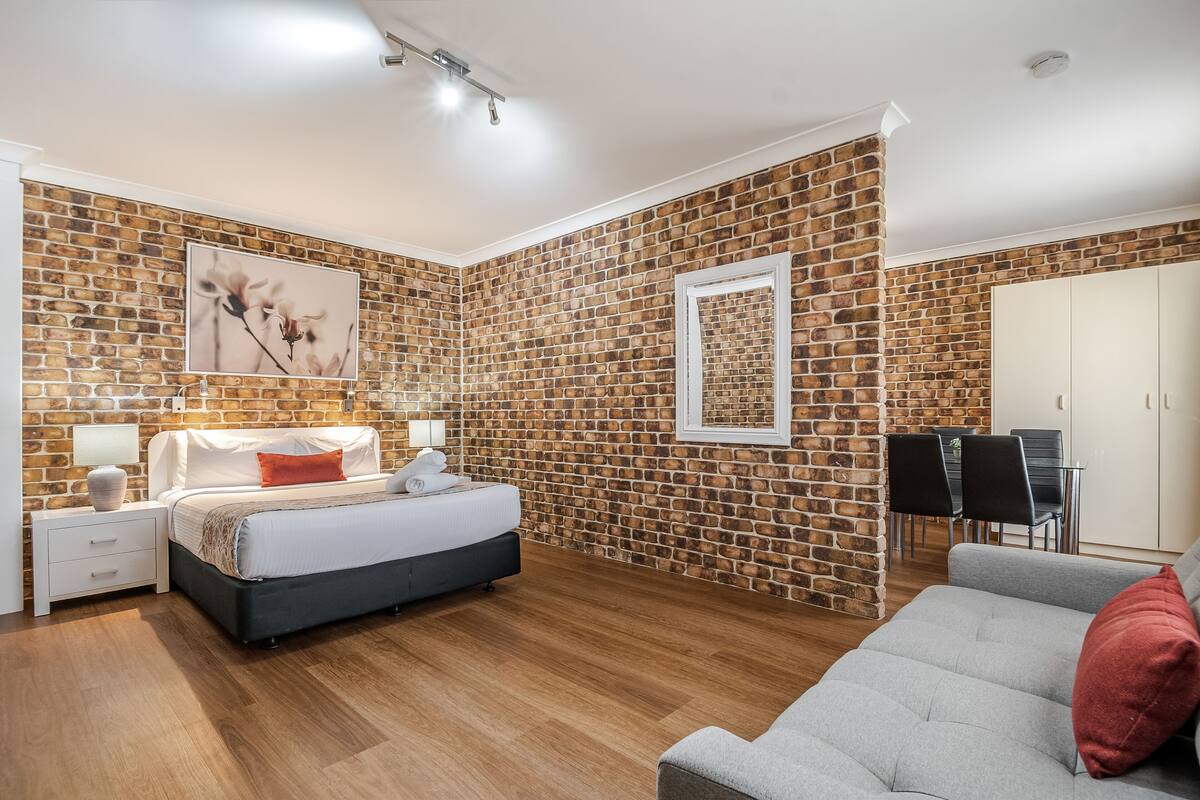 A bedroom with a brick wall, a bed, a nightstand, a lamp, a framed picture, a wardrobe, and a dining area with chairs and a table.