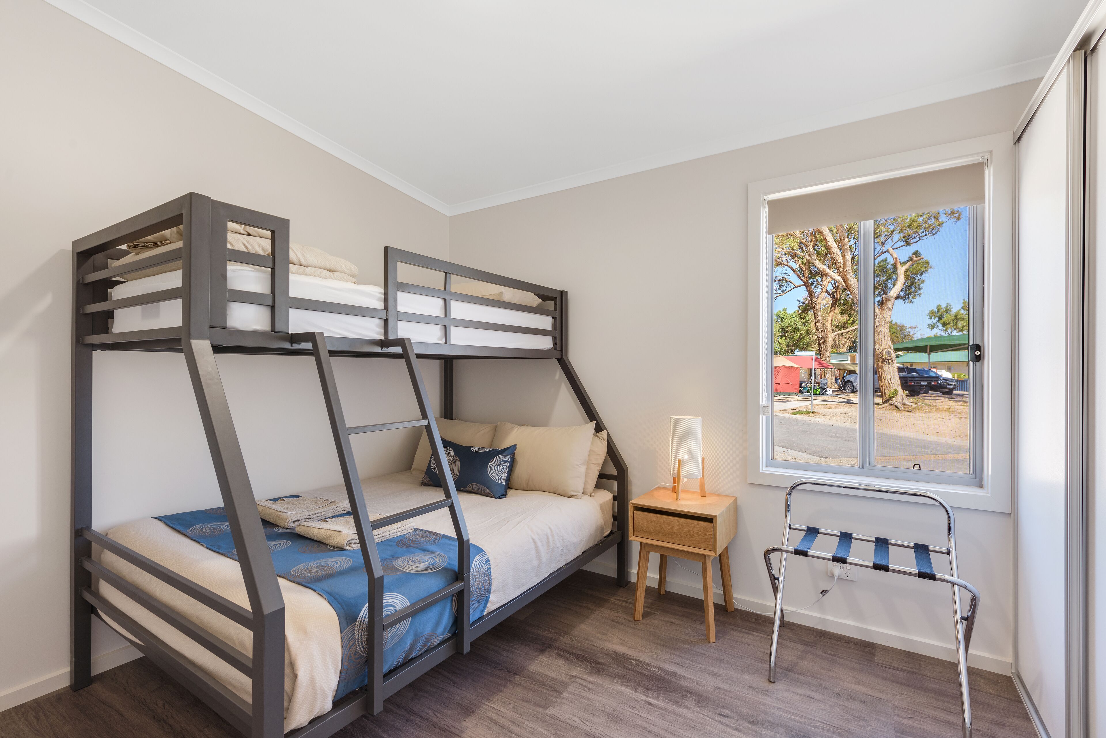 Une chambre avec un lit superposé donnant sur une fenêtre, une table de chevet avec une lampe et un étagère en métal.
