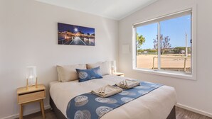 A bedroom with a large bed, a nightstand, a window with a view of the outdoors, and a painting on the wall. - Discovery Parks - Streaky Bay Foreshore (Streaky Bay)
