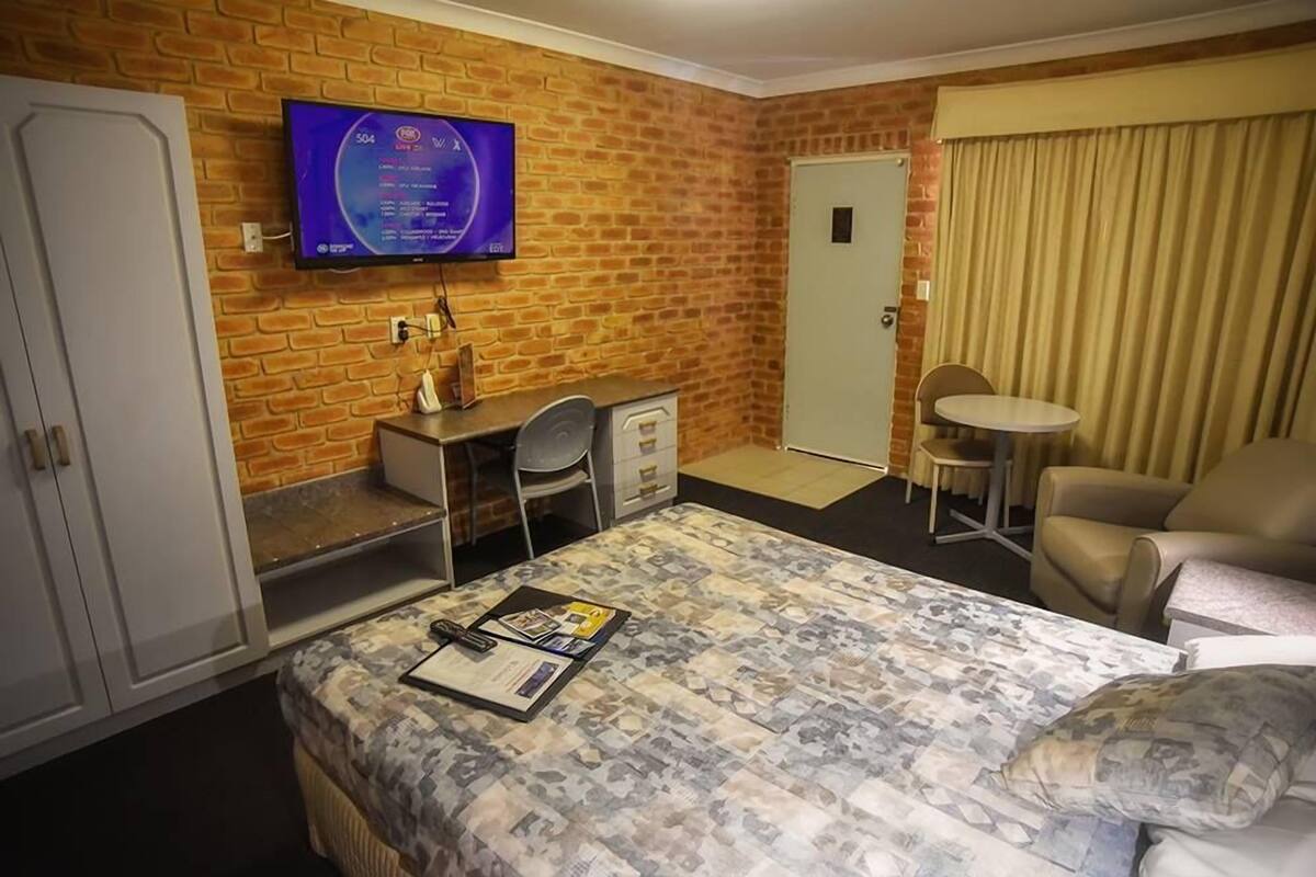 A hotel room with a brick wall, a TV mounted above a desk, a bed with a patterned bedspread, a chair, and a small table.